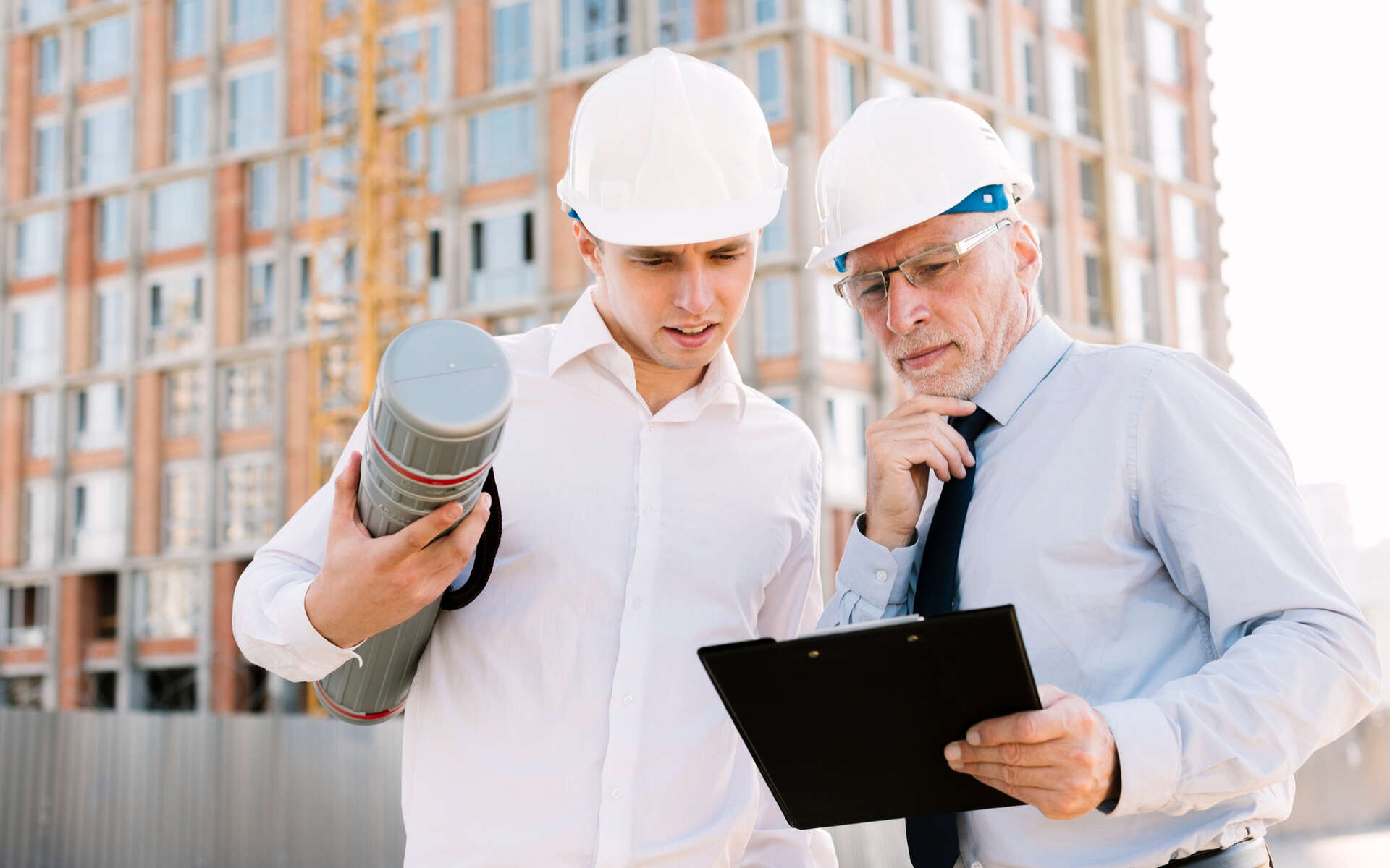 Two construction professionals in hard hats review documents on-site, discussing purchasing compliance for a building project.